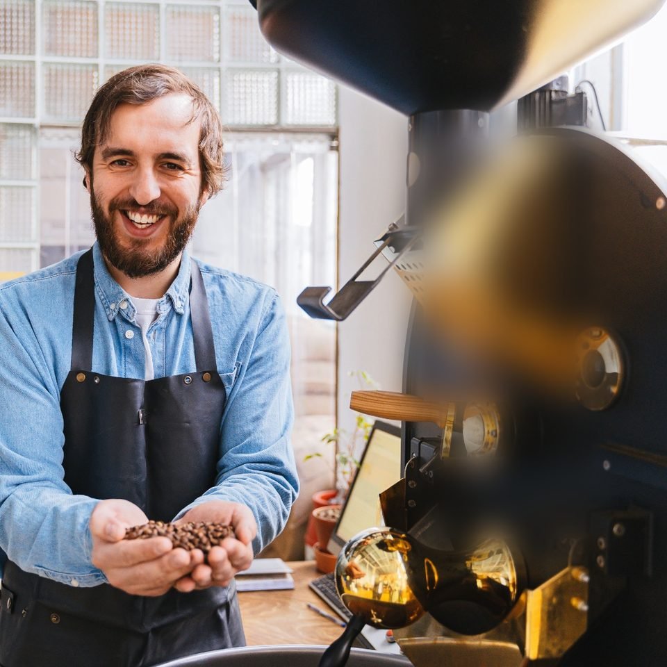 Röster mit Schürze steht lächelnd neben einer Trommelröstmaschine und hält frisch geröstete Kaffeebohnen in den Händen